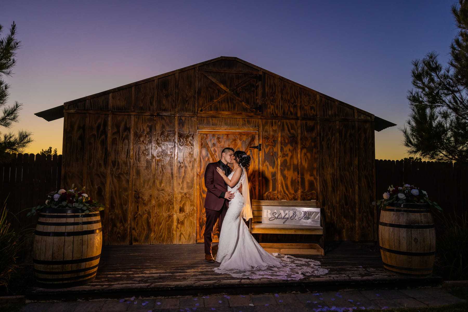 Bride and groom kissing in front of a rustic wooden barn with wine barrels at twilight