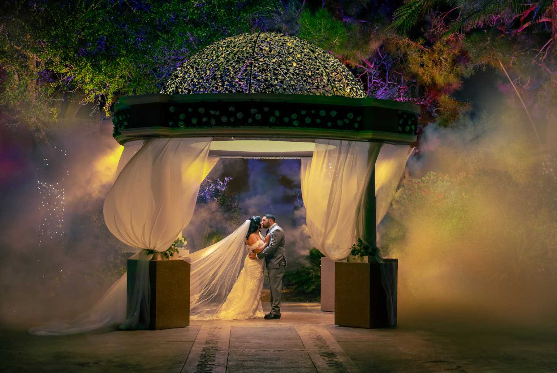 Bride and groom kissing under a garden gazebo with dramatic lighting and fog at Rainbow Gardens in Las Vegas