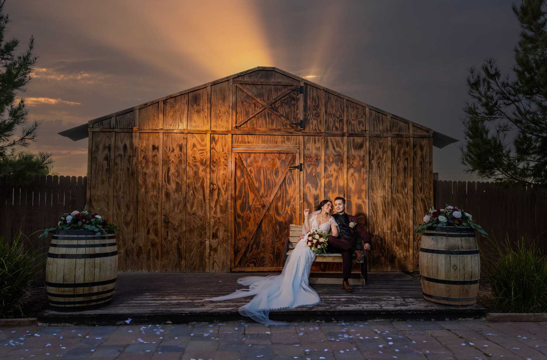 Bride and groom sitting in front of a rustic wooden barn with wine barrels and dramatic sunset sky