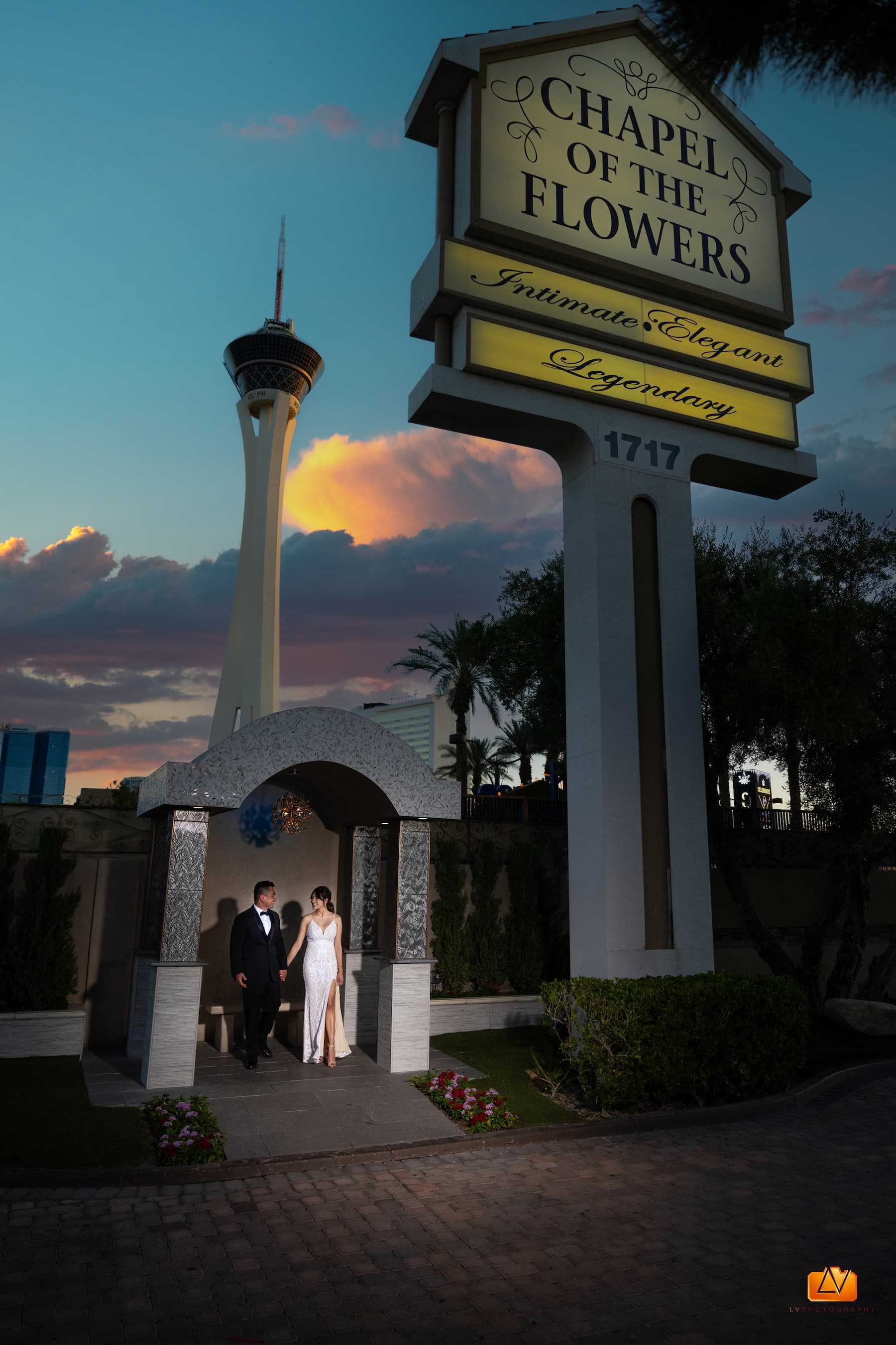 Bride and groom standing under the Chapel of the Flowers sign with the Las Vegas Strat tower in the background at dusk