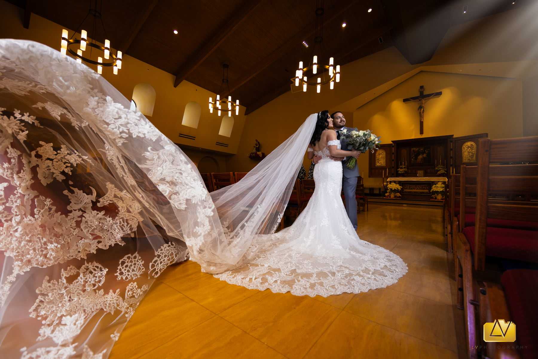 Bride and groom posing inside a Catholic church with the bride’s lace veil flowing across the aisle